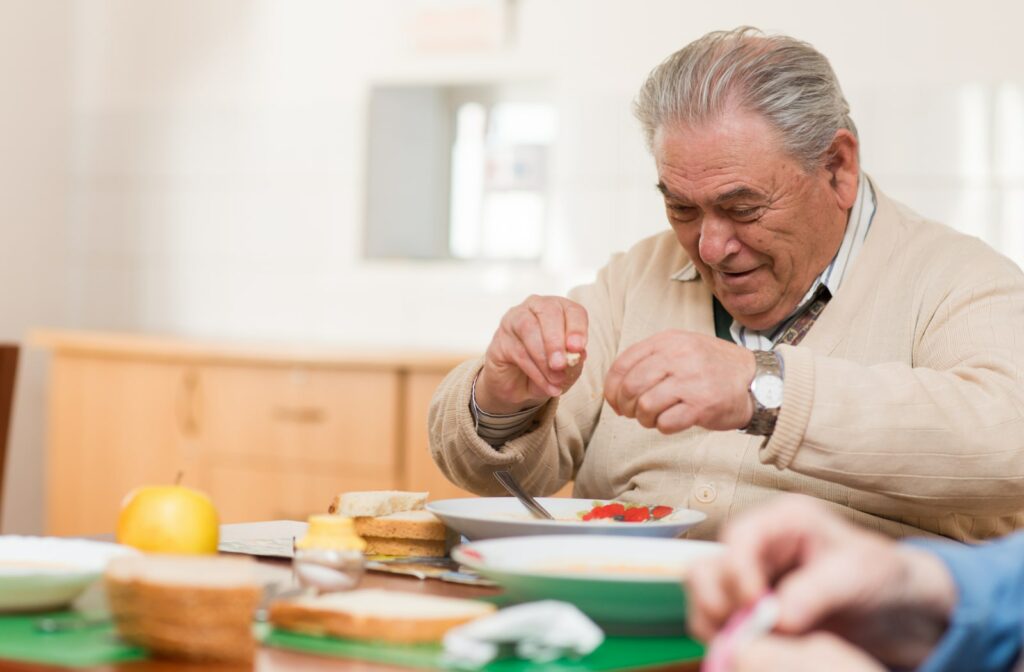 A mature smiling resident holds a small piece of bread in each hand as he looks down smiling at his lunch of soup and sandwich