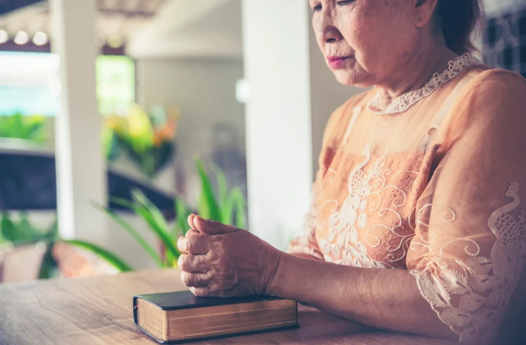 An older woman praying with her hands on her Bible.