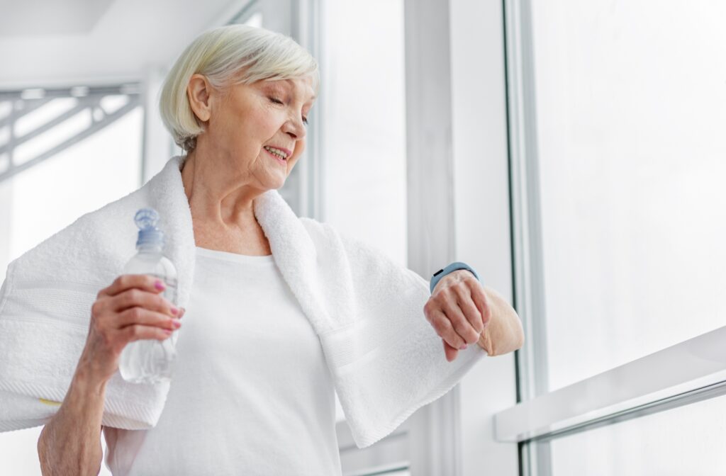 An older woman holding a bottle of water and a towel around her neck checks her fitness watch after a workout.