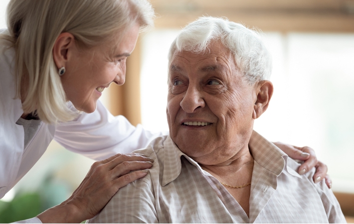 A smiling female caregiver smiles and holds the shoulders of a male resident as she checks in on him.