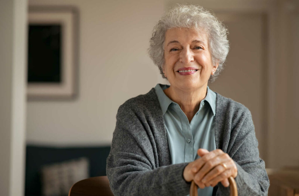 A senior woman with short grey hair is smiling and resting her hands on a cane as she looks directly at the camera.