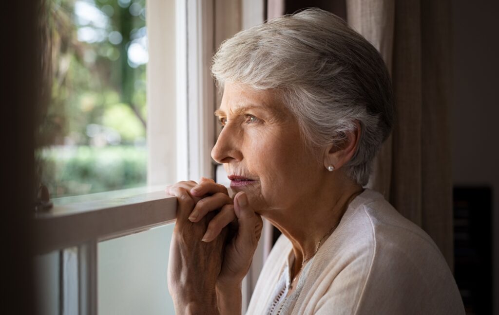 A senior stares thoughtfully out of an open window, with a green garden shown in the background.