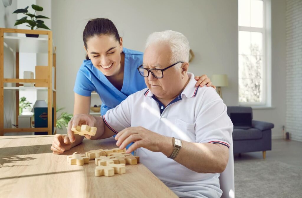 A caregiver smiles while helping a resident assemble a wooden block puzzle in the common room in memory care