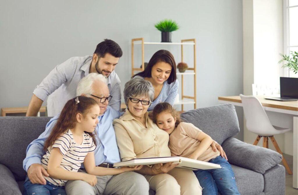 Two seniors sit on a sofa with their family while looking at photos together.