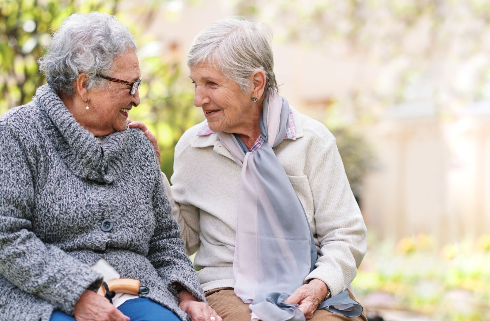 Two senior friends smile as they reminisce while sitting on a bench outdoors.