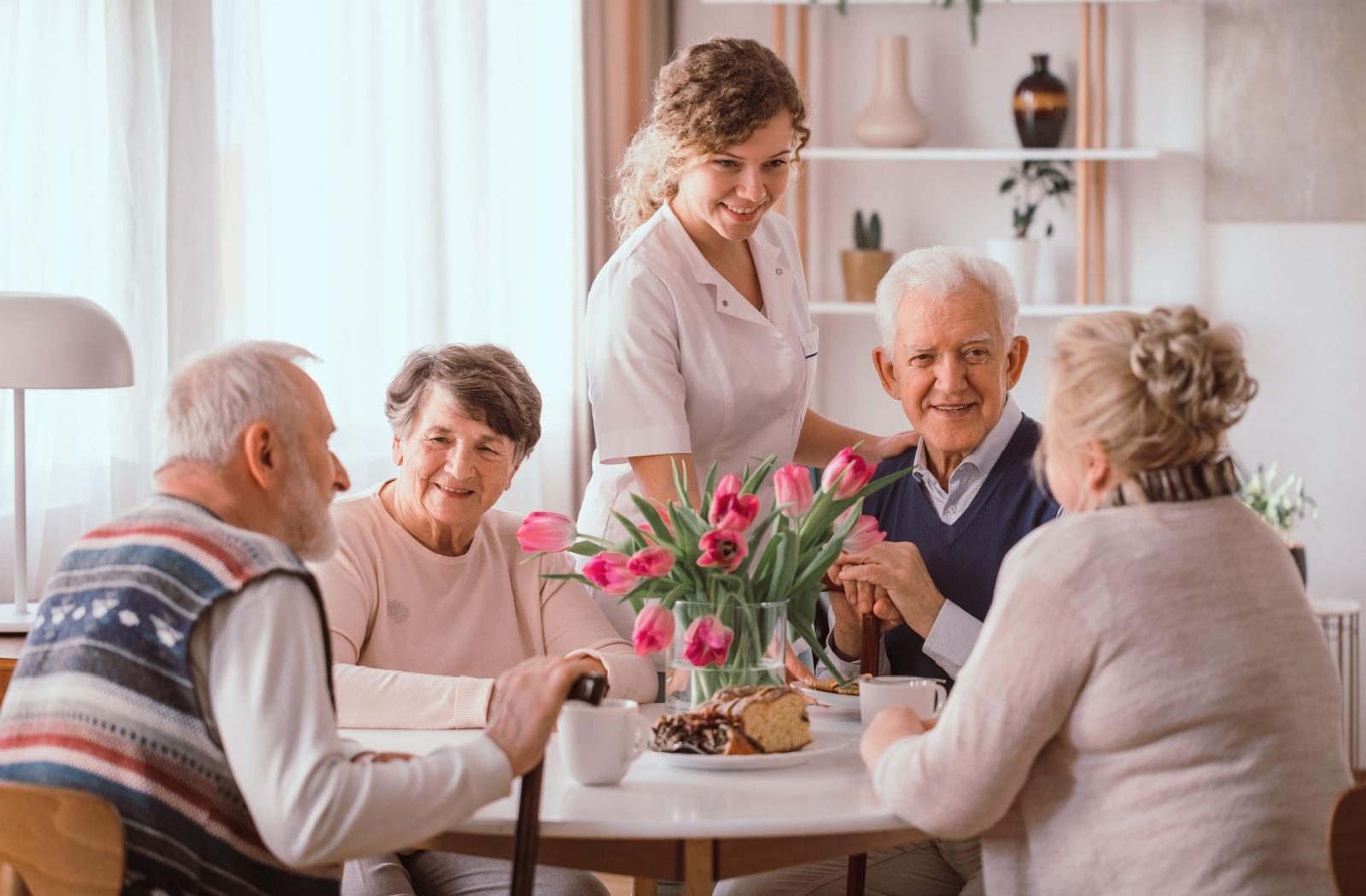 A group of senior friends enjoy a conversation over coffee, as a senior living community staff member serves food.
