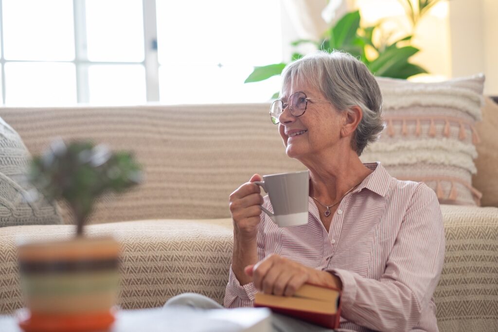 Senior adult woman smiling, sitting on the floor at senior living residence relaxing with a coffee cup and a book, enjoying independence